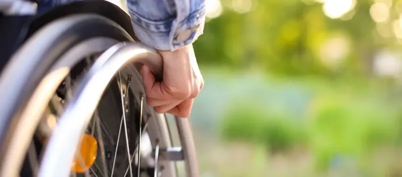Tekerlekli sandalyedeki bir kişinin eli, yeşil doğa fonunda destek arıyor.